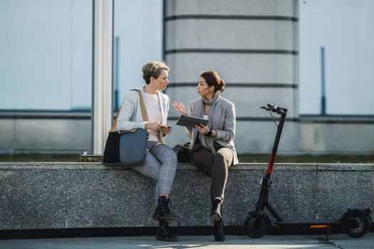 A two successful businesswomen using a digital tablet together while having a quick break in front of the office building.