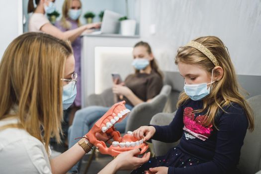 Female dentist and little girl wearing protective face masks while communicating in waiting room.