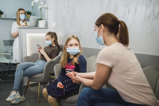 Worried little girl and her mom with face mask in waiting room at dentist's office.