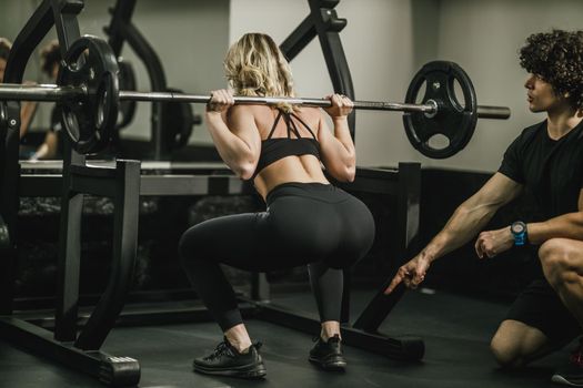 A young muscular woman is doing exercises for her legs supporting by coach in the gym.