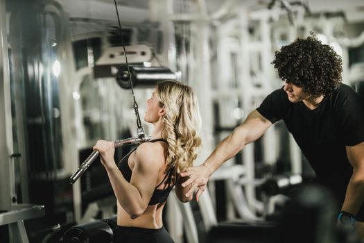 A young muscular woman is doing training at machine supporting by coach in the gym.