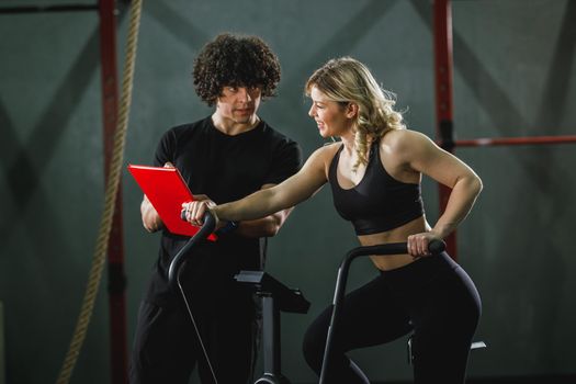 A young muscular woman is doing hard cal bike crossfit training in the gym supporting by coach.