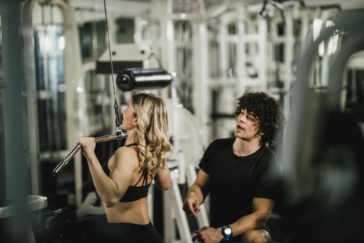 A young muscular woman is doing training at machine supporting by coach in the gym.