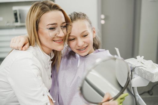 A happy teenager girl looking her teeth in a mirror after dental procedure at dentist's office.