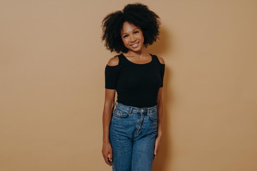 Indoor photo of pretty afro american smiling woman in casual outfit looking at camera with shy charming smile, posing isolated over dark beige background. Beautiful dark skinned women
