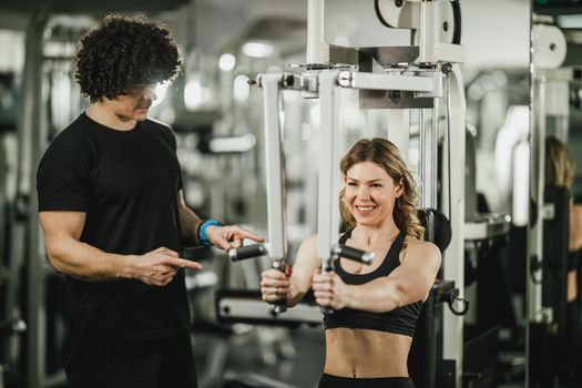 A young muscular woman is doing training at machine supporting by coach in the gym.