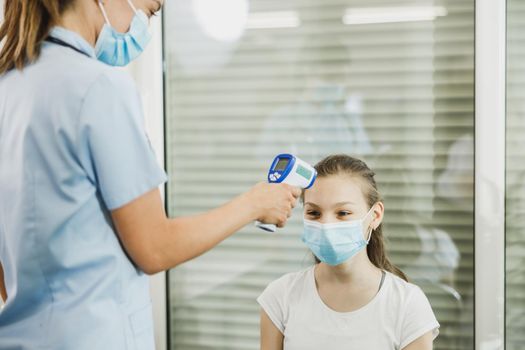 Nurse measuring temperature of a cute female teenager in waiting room during coronavirus pandemic.