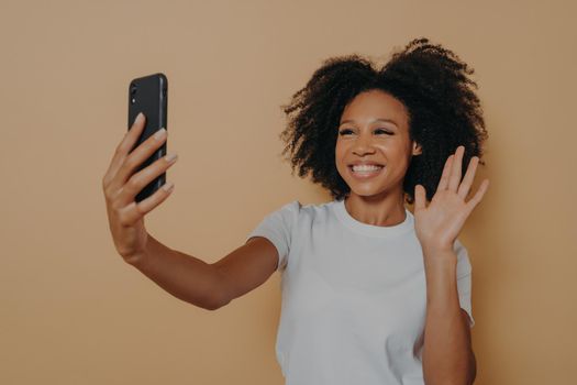 Hello! Beautiful cheerful dark skinned woman smiling at camera on modern smartphone, taking selfie on mobile phone and feeling happy, gesturing hi while making video call, isolated over studio wall