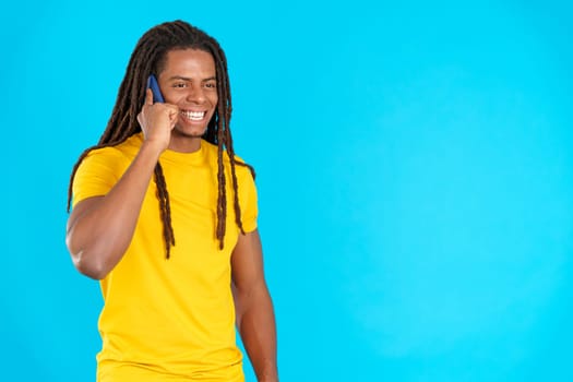 Latin man with dreadlocks smiling while talking to the mobile in studio with blue background