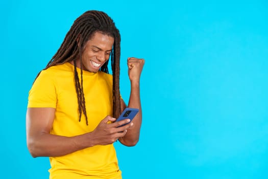 Excited latin man with dreadlocks celebrating while using the mobile phone in studio with blue background