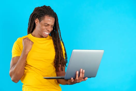 Latin man with dreadlocks celebrating while looking at the laptop in studio with blue background