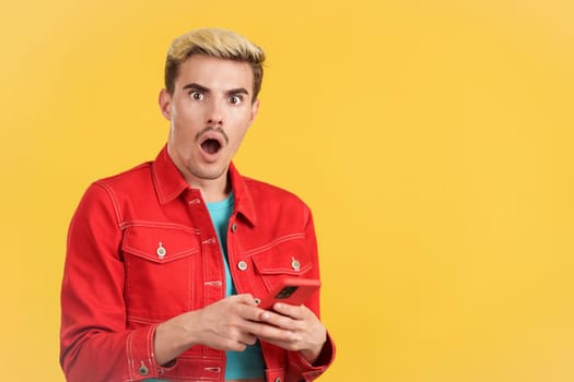 Astonished gay man looking the camera while using a mobile in studio with yellow background