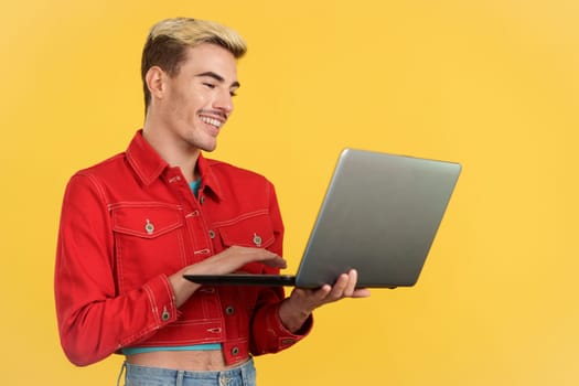 Fashionable gay man smiling while using a laptop in studio with yellow background