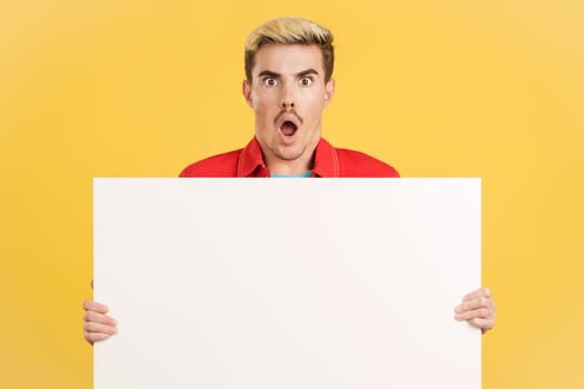 Amazed gay man holding a blank board in studio with yellow background