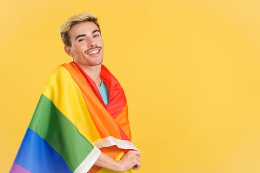 Happy gay man wrapped with a lgbt flag in studio with yellow background