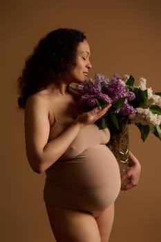 Attractive beautiful curly multiethnic gravid woman, pregnant expectant mother in beige lingerie, holding bouquet of blooming lilacs, isolated studio background. Pregnancy fashion. Body positivity