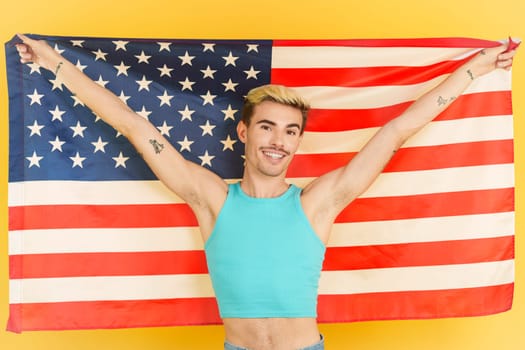 Happy gay man raising a north america national flag in studio with yellow background