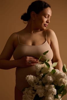 Pregnancy. Maternity. Expecting baby. Multiethnic attractive delightful young pregnant woman in beige lingerie, holding hand on her belly, posing with blooming white lilacs, isolated studio background