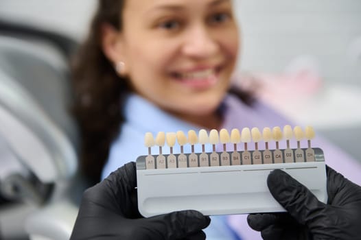 Selective focus on tooth color chart in the gloved hands of a doctor dentist, choosing the shade of veneers, according to Vita scale. Blurred smiling female patient at dental appointment in background