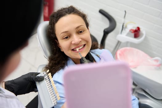 Close-up portrait of a smiling woman at dentist's appointment sitting in dental chair while a doctor dentist helping to pick teeth whitening shade from the chart, according to Vita scale color chart