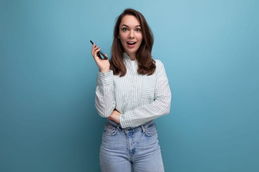 surprised young brunette lady dressed in a striped shirt and jeans tells the news holding a phone in her hand on a studio background.