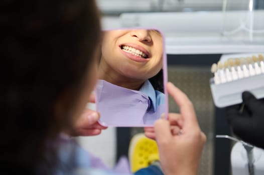 Beautiful smile of female patient, looking at her mirror reflection, after teeth bleaching procedure in dentistry clinic. Cropped view of dentist's hands hold a dental scale with teeth color and shade
