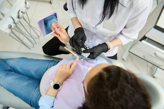 Overhead view of female patient in dentist chair discussing the color and shade of veneers with her orthodontist, pointing finger at the color and shade in the Vita scale color chart. Teeth prosthesis