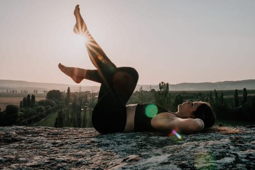 Well looking middle aged woman with long hair, fitness instructor in leggings and tops doing stretching and pilates on the rock near forest. Female fitness yoga routine concept. Healthy lifestyle.