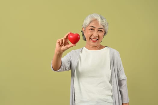 Beautiful senior woman holding a red heart and smiling to camera. Cardiology, medical and healthcare concept.