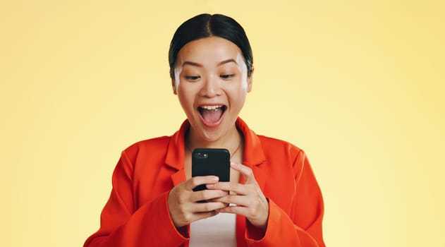 Winner, success celebration and Asian woman with phone in studio isolated on a yellow background. Surprise, fist pump or happy female with mobile to celebrate after winning lottery prize or good news.