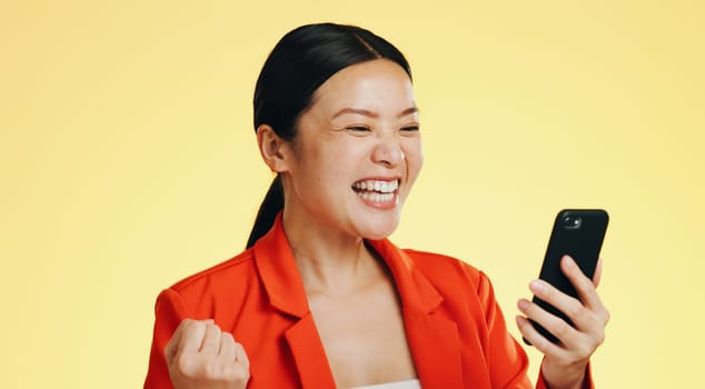 Winner, success celebration and Asian woman with phone in studio isolated on a yellow background. Surprise, fist pump or happy female with mobile to celebrate after winning lottery prize or good news.