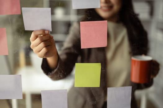 Smiling businesswoman executive manager reading sticky notes on transparent board, planning goals, manage corporate work tasks.