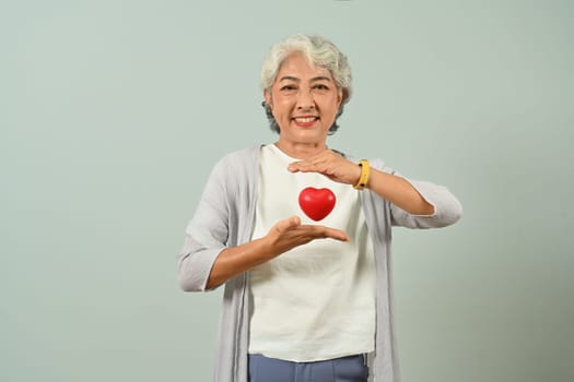 Smiling 60s woman holding red heart over blue background. Health care, medical and charity concept.