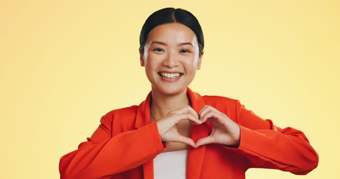 Smile, heart in hands and face of Asian woman on yellow background for love, support and charity mockup. Emoji, hand gesture and portrait of happy girl in studio with shape for care, trust and kind.