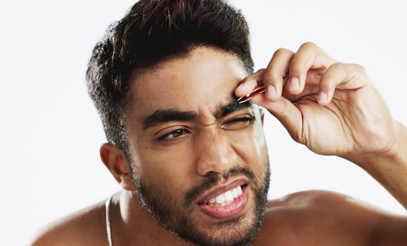 Man grooming his eyebrows with a tweezer in studio for self care, beauty and cleanliness. Hair removal, tweezing and male model from India doing facial epilation plucking routine by white background