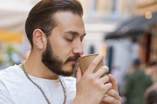 Close-up of happy bearded young man enjoying morning coffee hot drink and smiling outdoors. Relaxing, taking a break. Guy walking in urban city sunshine street, drinking coffee to go. Town lifestyles