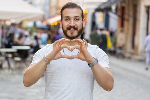 I love you. Young man makes symbol of love, showing heart sign to camera, express romantic feelings, express sincere positive feelings. Charity, gratitude, donation. Outdoors in urban city street