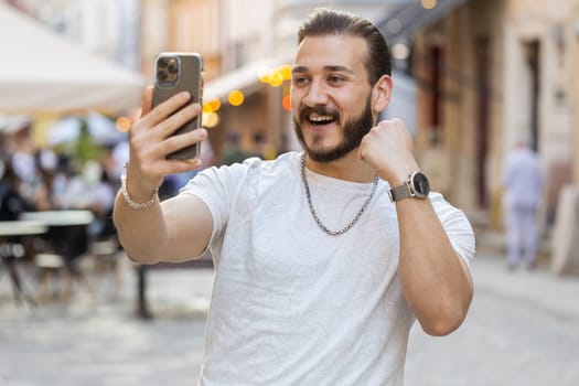 Bearded young man use mobile smartphone celebrating win good message news, lottery jackpot victory, giveaway online outdoors. Happy guy tourist walking in urban sunshine city street. Town lifestyles