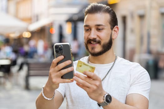 Young man using credit bank card smartphone while transferring money, purchases online shopping, order food delivery, booking hotel room. Guy tourist walking in urban sunshine city street outdoors
