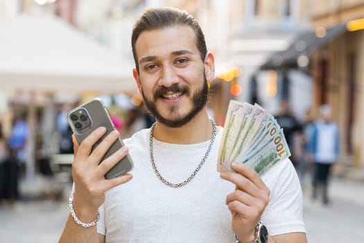 Happy rich young man counting money dollar cash, use smartphone calculator app, plans to order gifts and food delivery online, booking hotel room. Bearded guy tourist in city street. Town outdoors