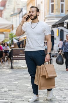 Joyful smiling bearded young man shopaholic consumer after shopping sale with full bags with gifts outdoors. Guy tourist traveler walking along the urban sunshine city street road. Town lifestyles