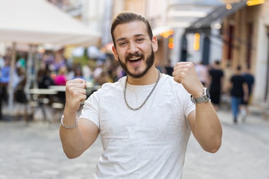 Portrait of bearded young man shouting, celebrating success, winning, goal achievement, good news, lottery jackpot luck, victory outdoors. Guy walking in urban city sunshine street. Town lifestyles