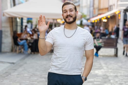 Portrait of bearded young man smiling friendly at camera, waving hands gesturing hello, hi, greeting or goodbye, welcoming with hospitable expression outdoors. Guy walking in urban city town street