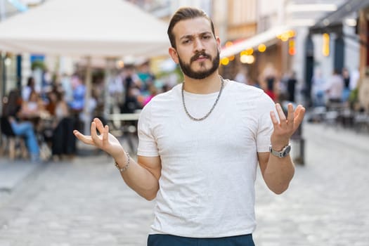 What. Why. Surprised frustrated bearded young man raising hands asking reason of failure, demonstrating disbelief irritation by troubles outdoors. Confused guy walking in urban city sunshine street