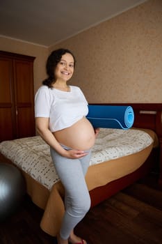 Full length portrait multi ethnic positive pregnant woman, putting her hand on her big belly in 28 week of pregnancy, smiling looking at camera, ready for prenatal fitness and stretching exercises