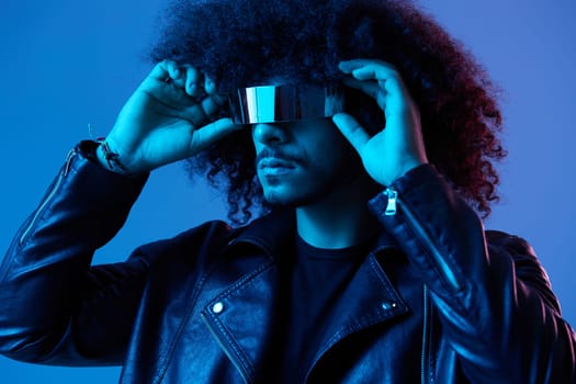 Portrait of fashion man with curly hair with stylish glasses on blue background multinational, colored light, black leather jacket trend, modern concept. High quality photo