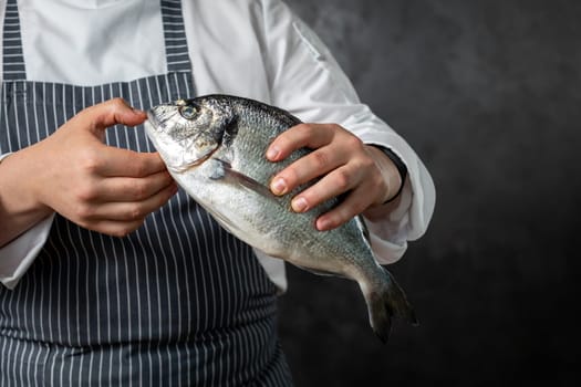 Crop anonymous male fishmonger in striped apron holding fresh uncooked fish against grungy black background