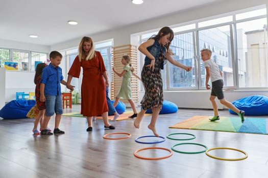 Small nursery school children with female teacher on floor indoors in classroom, doing exercise. Jumping over hula hoop circles track on the floor