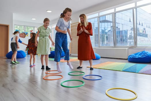 Small nursery school children with female teacher on floor indoors in classroom, doing exercise. Jumping over hula hoop circles track on the floor