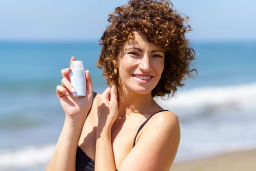 Happy curly haired female in black bikini standing near waving sea on sunny day and demonstrating blank bottle of sunscreen